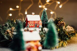 This image features a tin/metal house ornament that has white snow on the roof. The ornament is surrounded by small artificial christmas trees and is shown in keeping with our blog post about getting your home roof ready for the holidays