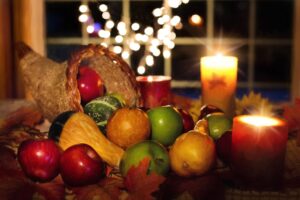 This is a bright and cheerful holiday image that shows a cornucopia (horn-shaped basket) with brightly colored fruit (red and green apples) and green and yellow squash spilling out onto a table with red and yellow light candles and a string of white twinkle lights hanging near the window in the background.